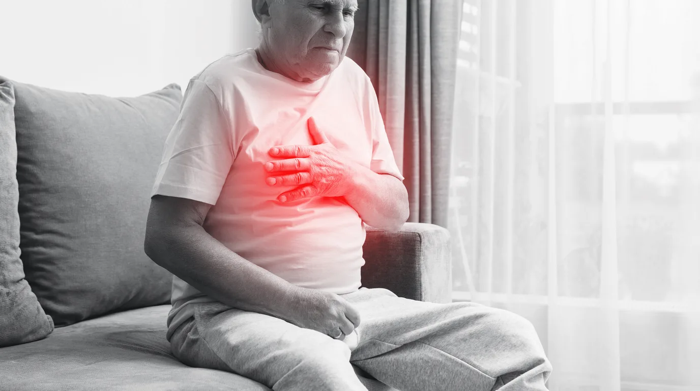 Older adult holding the center of the chest while seated at home, a common presentation of cardiac chest pain